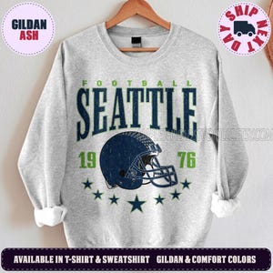 May include: A light grey crewneck sweatshirt featuring a dark blue and green graphic print. The design includes the text "FOOTBALL SEATTLE 19 76" above a dark blue football helmet illustration, accented by a series of small dark blue stars.