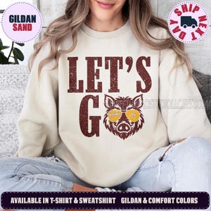 May include: A sand-colored crewneck sweatshirt featuring the dark red distressed text LET'S GO above a graphic of a wild boar wearing yellow sunglasses. The sunglasses lenses contain the text WASHINGTON. The design is centered on the chest.