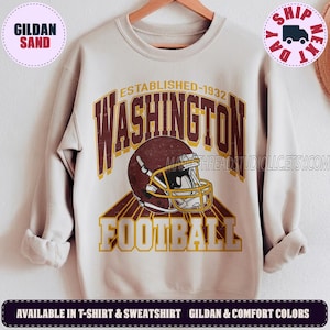 May include: A sand-colored crewneck sweatshirt featuring a graphic print with the text "WASHINGTON" in dark red, "ESTABLISHED-1932" in yellow, and "FOOTBALL" in yellow. A dark red football helmet with a yellow face mask is centered in the design.