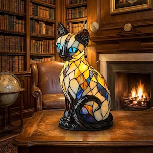 Op de afbeelding: Een glas-in-loodlamp in de vorm van een zittende kat. De lamp heeft een mozaïekpatroon van gele, amberkleurige en blauwe glasstukjes met zwarte omlijningen. De kat heeft fel oplichtende blauwe ogen en een zwart gezicht, oren en een zwarte staart.