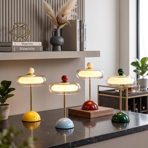 May include: Four table lamps with thin gold-tone metal stems, circular bases in yellow, light blue, red, and dark green, and clear glass shades containing glowing white light diffusers topped with small matching spherical finials.