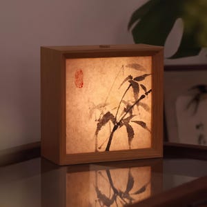 Pode incluir: Uma luminária quadrada de madeira com uma moldura de cor clara e um painel translúcido com uma pintura a tinta preta de caules e folhas de bambu. Um pequeno selo vermelho é visível no canto superior esquerdo da superfície de papel.