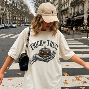 Può includere: Una t-shirt color crema con la grafica di una mano scheletrica che regge una zucca luminosa con la scritta TRICK OR TREAT sopra, abbinata a un berretto da baseball beige e una borsa a tracolla nera trapuntata con catena dorata.