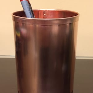 Peut inclure: Un organiseur de bureau en cuivre avec une finition lisse et brillante. Deux stylos dépassent du haut de l'organisateur.