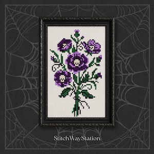 May include: A framed cross-stitch embroidery featuring a bouquet of purple flowers with small white skulls at their centers, set against a white background. The artwork is enclosed in a black rectangular frame with a decorative border.