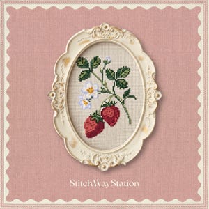 Puede incluir: Un marco ovalado de color crema con adornos que contiene un bordado de punto de cruz de dos fresas rojas, hojas verdes y pequeñas flores blancas sobre un fondo de tela beige claro. El texto StitchWayStation aparece debajo del marco.