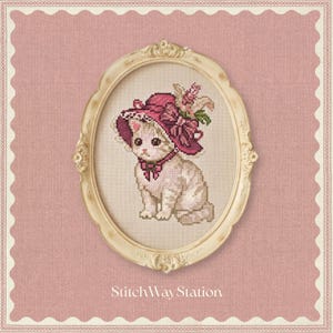 Puede incluir: Un bordado de punto de cruz de un gatito de color claro que lleva un gorro rosa oscuro con un lazo y detalles florales, enmarcado en un marco ovalado, ornamentado y de color crema. El texto StitchWay Station está impreso en blanco abajo.