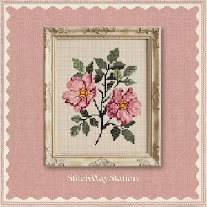 May include: A cross-stitch embroidery of two pink flowers with yellow centers and green leaves on a cream-colored fabric background, encased in an ornate, distressed off-white wooden frame. The text StitchWayStation appears below the frame.