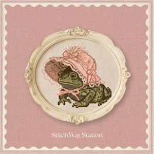 Puede incluir: Un bordado de punto de cruz de una rana verde con un gorro rosa, enmarcado en un marco ovalado de color crema. El diseño se presenta sobre un fondo rosa texturizado con un borde festoneado. El texto StitchWayStation aparece debajo.