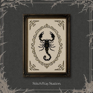 Puede incluir: Un bordado en punto de cruz enmarcado que presenta un escorpión negro centrado dentro de un borde ovalado verde oscuro ornamentado sobre un fondo de tela color crema. El diseño incluye detalles decorativos en las esquinas y el texto StitchWayStation.