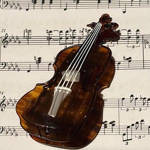 May include: A violin-shaped brooch or accessory crafted from translucent, dark brown tortoiseshell-patterned material. It features white, raised lines representing the strings, a bridge, and a fingerboard, with a detailed scroll at the top.