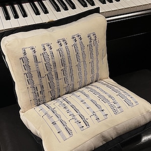 May include: A rectangular cream-coloured cushion featuring a black printed musical score with the text Golden Hour. The cushion has a black border and is positioned upright, displaying two pages of sheet music with detailed staves and notes.
