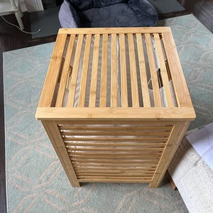May include: A square laundry hamper constructed from light-toned bamboo slats. The hamper features a slatted lid and sides with integrated handles, revealing a white fabric liner inside. The structure is composed of horizontal and vertical bamboo pieces.
