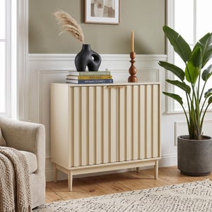 May include: A cream-colored cabinet with vertical fluted doors and thin gold handles, standing on four tapered legs. On top sits a black abstract vase with pampas grass, a stack of three books, and a tall brown wooden candle holder.