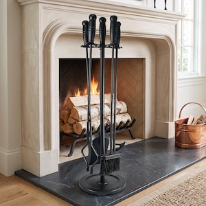 May include: A black fireplace tool set featuring a circular base and a central stand with five hanging implements. The set includes a poker, a shovel, a brush with black bristles, and tongs, all finished in a matte black metal with rounded handles.