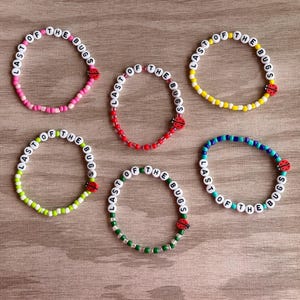May include: Five circular beaded bracelets, each featuring white letter beads spelling out the phrase LAST OF THE BUGS and a single red ladybug charm. The bracelets are made with colourful small beads in pink, red, yellow, green, and blue.