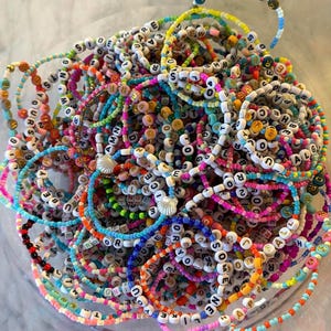 May include: A collection of colorful beaded bracelets featuring white letter beads with black text, interspersed with small multi-colored seed beads. Some bracelets include small white seashell charms, creating a vibrant, layered accessory set.