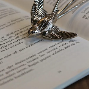 May include: A polished silver-toned metal swallow bird brooch with detailed wing and tail feather engravings. The bird is depicted in a mid-flight pose with wings spread upward and a long, forked tail, featuring a reflective, smooth metallic finish.