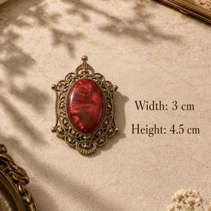 Könnte beinhalten: Eine ovale Brosche mit einem tiefroten, marmorierten Cabochon, gefasst in einen aufwendigen Rahmen aus antik bronzefarbenem Metall. Das Stück ist 3 cm breit und 4,5 cm hoch und zeigt detaillierte Filigranarbeiten um den zentralen Stein.