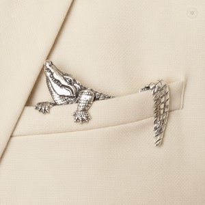 May include: A silver-toned metal brooch shaped like an alligator, designed to appear as if the creature is crawling out of a pocket. The piece features detailed scales, a textured surface, and a separate tail section that rests on the pocket edge.