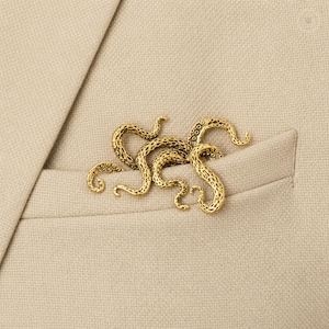 Puede incluir: Un broche de metal dorado con forma de grupo de tentáculos de pulpo con texturas detalladas de ventosas. El broche está sujeto a la tela de una americana beige y presenta múltiples brazos curvos que se extienden desde el punto central.