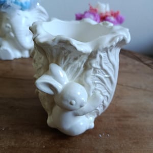 Peut inclure: Un cache-pot en céramique blanche en forme de feuille de chou avec une petite figurine de lapin fixée sur le devant. La surface présente une finition brillante et des nervures en relief détaillées sur toute la structure.