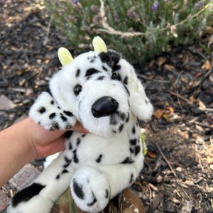 May include: A plush stuffed animal designed to look like a dalmatian dog with white fur and black spots. The toy features two small, pale yellow horns on top of its head, dark plastic eyes, and a black nose. It has floppy ears and soft paws.