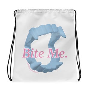Puede incluir: Una bolsa de cordón blanca con un gráfico azul claro de colmillos de vampiro de plástico dispuestos en forma circular. El texto "Bite Me." está impreso en rosa en el centro, con "Certified Nostalgia Co." en texto rosa más pequeño debajo.