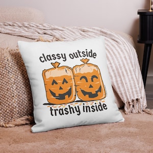 May include: A white square throw pillow featuring a graphic of two orange, jack-o'-lantern-style bread loaves in plastic bags. The text "classy outside" appears above the loaves, and "trashy inside" is printed below in black, bold lettering.
