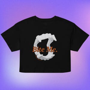 May include: A black cropped t-shirt featuring a graphic of white vampire fangs arranged in a circular shape. The text "Bite Me." is printed in orange across the center of the fangs, with smaller text below reading "Cryptid Starlight Co."