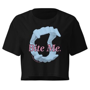 May include: A black cropped t-shirt featuring a graphic of light blue vampire fangs arranged in a circular shape. The text "Bite Me." is printed in pink across the center of the fangs, with the small text "Certified Nostalgia Co." below.