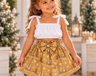 May include: A two-piece outfit consisting of a white sleeveless crop top with shoulder ties and a mustard yellow skirt featuring a repeating pattern of white snowflakes and reindeer-drawn sleighs, finished with a central bow at the waist.