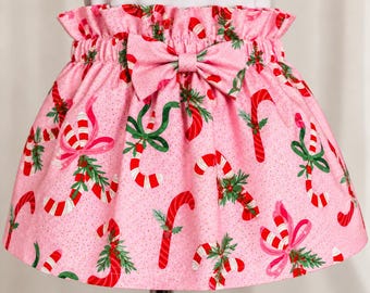 May include: A pink skirt featuring a repeating pattern of red and white candy canes, green holly leaves, and pink bows. The skirt has a gathered, elasticized paper-bag waist with a matching fabric bow centered at the front.