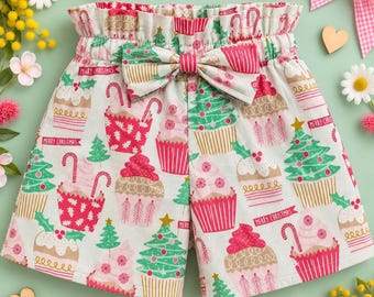 May include: High-waisted shorts featuring a festive print of cupcakes and Christmas trees in red, green, and tan on a white background. The shorts include a matching fabric bow at the center of the elasticized, ruffled waistband.