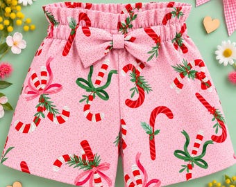 May include: Pink shorts featuring a repeating pattern of red and white candy canes, green pine sprigs, and pink bows. The shorts have a high-waisted, elasticized paper bag style waistband with a matching fabric bow centered at the front.