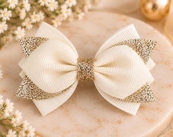 May include: A hair bow constructed from white ribbed grosgrain ribbon, featuring layered gold glitter accents on the outer edges and a central gold glitter knot. The bow is symmetrical with a structured, multi-layered design.