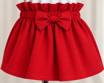 May include: A bright red skirt featuring a high-waisted paper bag design with an elasticized waistband and a decorative fabric bow centered at the front. The skirt has a flared silhouette and a textured finish, worn over a plain white top.