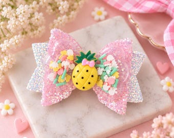 May include: A hair bow featuring a pink glittery base and a clear center section filled with colorful sprinkles and small shapes. A yellow pineapple charm with a small pink bow sits in the middle of the bow, which is layered with a white glittery backing.