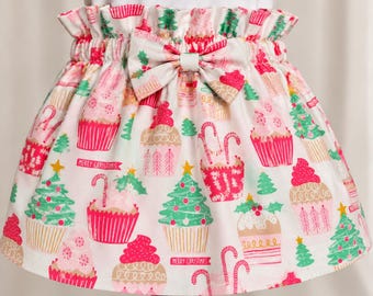 May include: A white skirt with a ruffled elastic waistband and a central bow. The fabric features a repeating pattern of pink and red cupcakes, green Christmas trees, and candy canes, with the text Merry Christmas printed on banners.