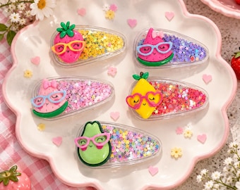 May include: Five clear triangular hair clips filled with colorful star-shaped glitter. Each clip features a 3D fruit character: a pink strawberry, two pink watermelon slices, a yellow lemon, and a green avocado, all wearing bright plastic glasses.