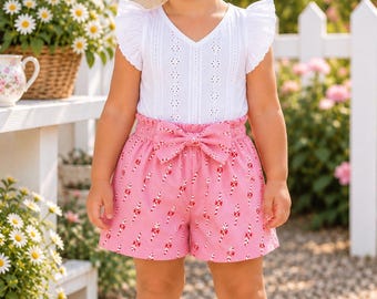 May include: A white sleeveless top with eyelet detailing and ruffled sleeves paired with pink shorts featuring a candy cane print and a front bow. The outfit includes a matching white hair bow and white sandals with a floral cutout pattern.
