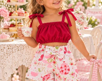 May include: A two-piece outfit consisting of a solid dark pink sleeveless crop top with shoulder tie straps and white shorts featuring a colorful pattern of pink teapots, strawberries, flowers, and small birds with a bow at the waist.