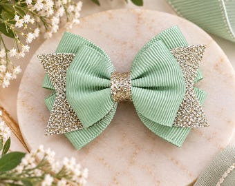May include: A hair bow constructed from sage green ribbed grosgrain ribbon, featuring layered sections accented with gold glitter fabric on the outer edges and a central gold glitter knot, creating a multi-dimensional appearance.
