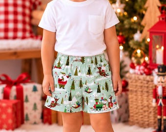 May include: A white short-sleeved t-shirt with a chest pocket paired with light green shorts featuring a festive holiday print of Santa Claus, reindeer, Christmas trees, and a train. The outfit is completed with simple white sneakers.
