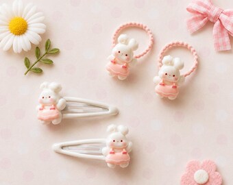 May include: Two white metal hair clips and two pink elastic hair ties, each featuring a small white rabbit figurine wearing a pink dress. The set includes a white daisy flower and a pink gingham fabric bow as decorative accents.