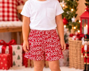 May include: A white short-sleeve t-shirt with a chest pocket paired with red shorts featuring a repeating white text pattern that reads HO HO HO. The outfit is completed with simple white sneakers, creating a clean and festive look.