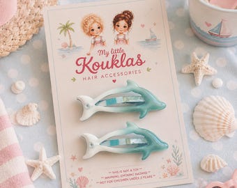 May include: Two dolphin-shaped hair clips on a display card labeled My little Kouklas Hair Accessories. The clips feature a gradient color transition from white at the tail to teal at the head. The card includes illustrations of two children, a palm tree, and a sailboat, with safety warnings printed at the bottom.