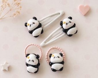 May include: Two white metal snap hair clips and two pink elastic hair ties, each featuring a small, glossy, three-dimensional panda bear figurine. Each panda has black and white coloring, a tiny yellow flower detail, and a smooth, rounded finish.