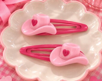 May include: Two hair clips featuring a light pink cowboy hat ornament with a small dark pink heart detail on the front. The clips themselves are a solid dark pink color and have a classic snap-style design for securing hair in place.