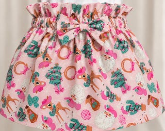 May include: A pink skirt with an elasticized ruffled waistband and a matching fabric bow. The fabric features a repeating pattern of Santa Claus figures, deer, Christmas trees, cacti, and disco balls in shades of pink, green, and brown.